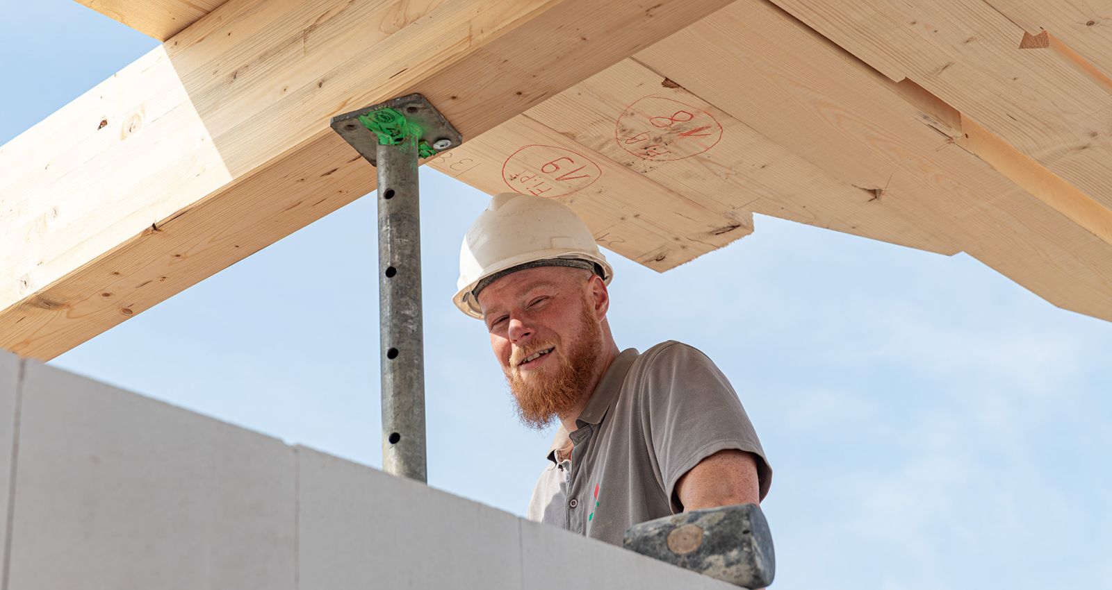 Ein fröhlicher Mitarbeiter unseres Bauhandwerkes bei Rohbauarbeiten an einem Dachgeschoss, mit Bauhelm und T-Shirt unter blauem Himmel.