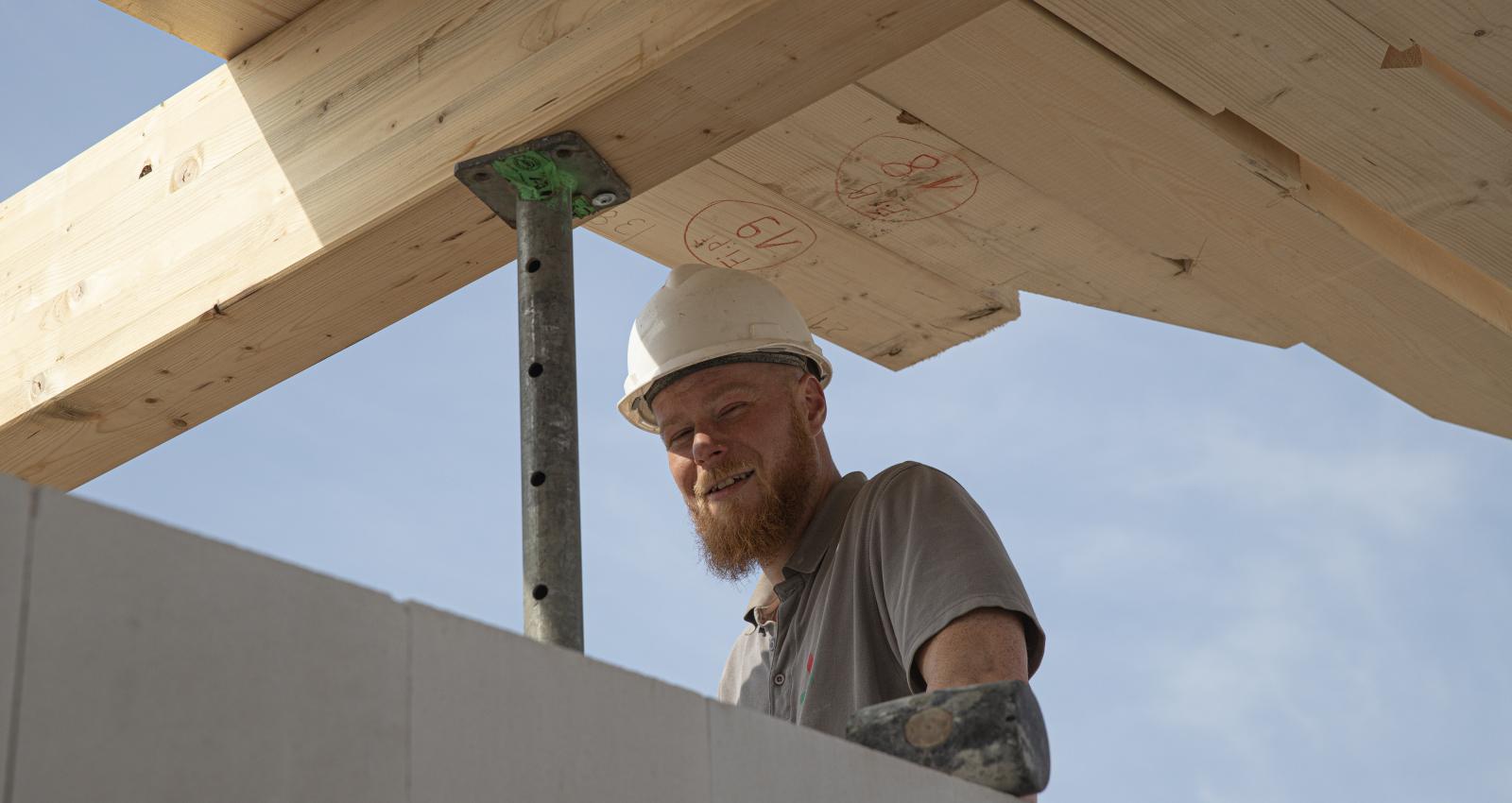 Ein fröhlicher Mitarbeiter unseres Bauhandwerkes bei Rohbauarbeiten an einem Dachgeschoss, mit Bauhelm und T-Shirt unter blauem Himmel.
