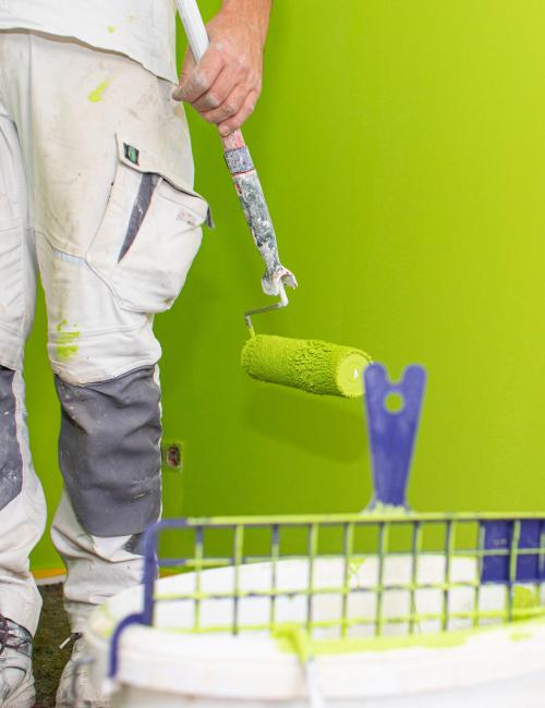 Maler bei Renovierungsarbeiten mit Malerrolle an Farbeimer, vor grüner, abgeklebter Wand