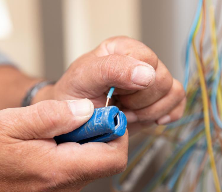 Bildausschnitt mit zwei Händen eines Elektromeisters beim abisolieren eines blauen Kabels mittels Abisolierzange.