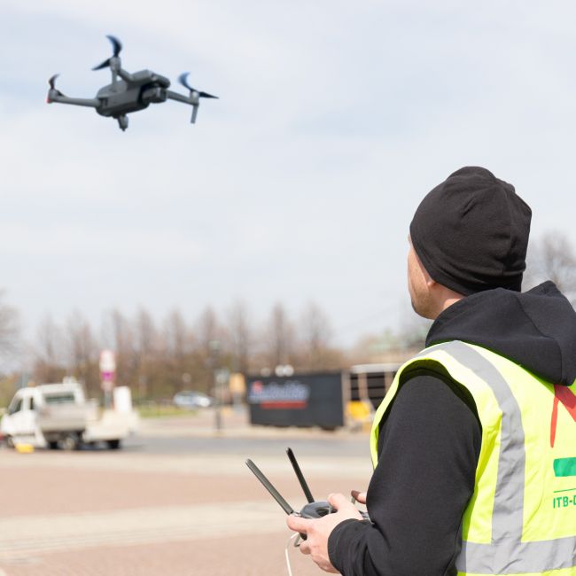 Aufsteigende Drohne bei bewölktem Himmel fliegt über eine Freifläche. Im Vordergrund zu sehen ist er Drohnenpilot der ITB-Dresden GmbH mit der Steuerung in der Hand