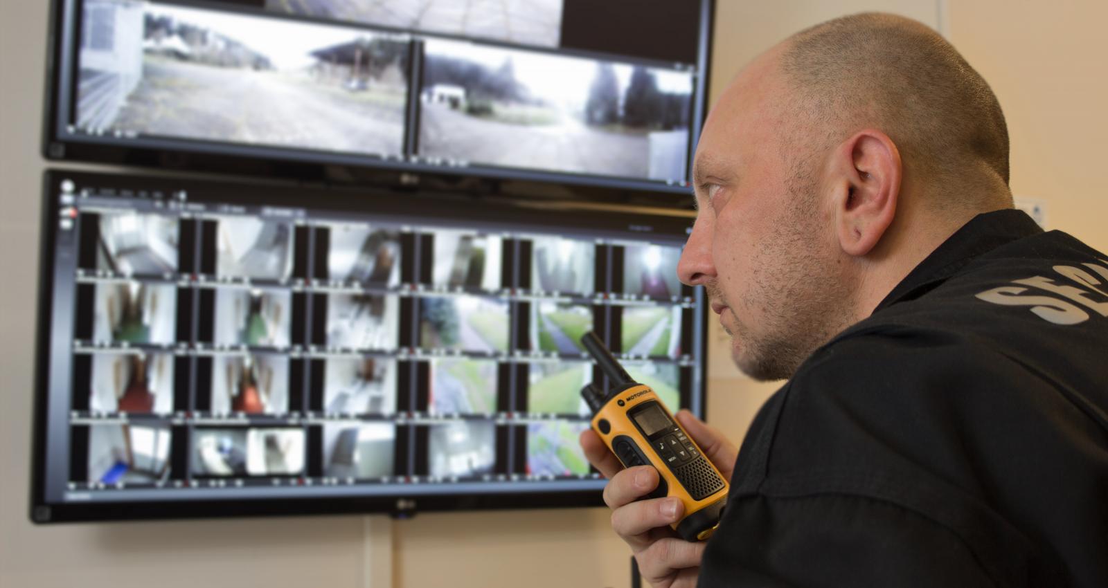 Ein Wachmann mit walkie talkie in der Hand überprüft an den Monitoren die Übertragung der Überwachungskameras.