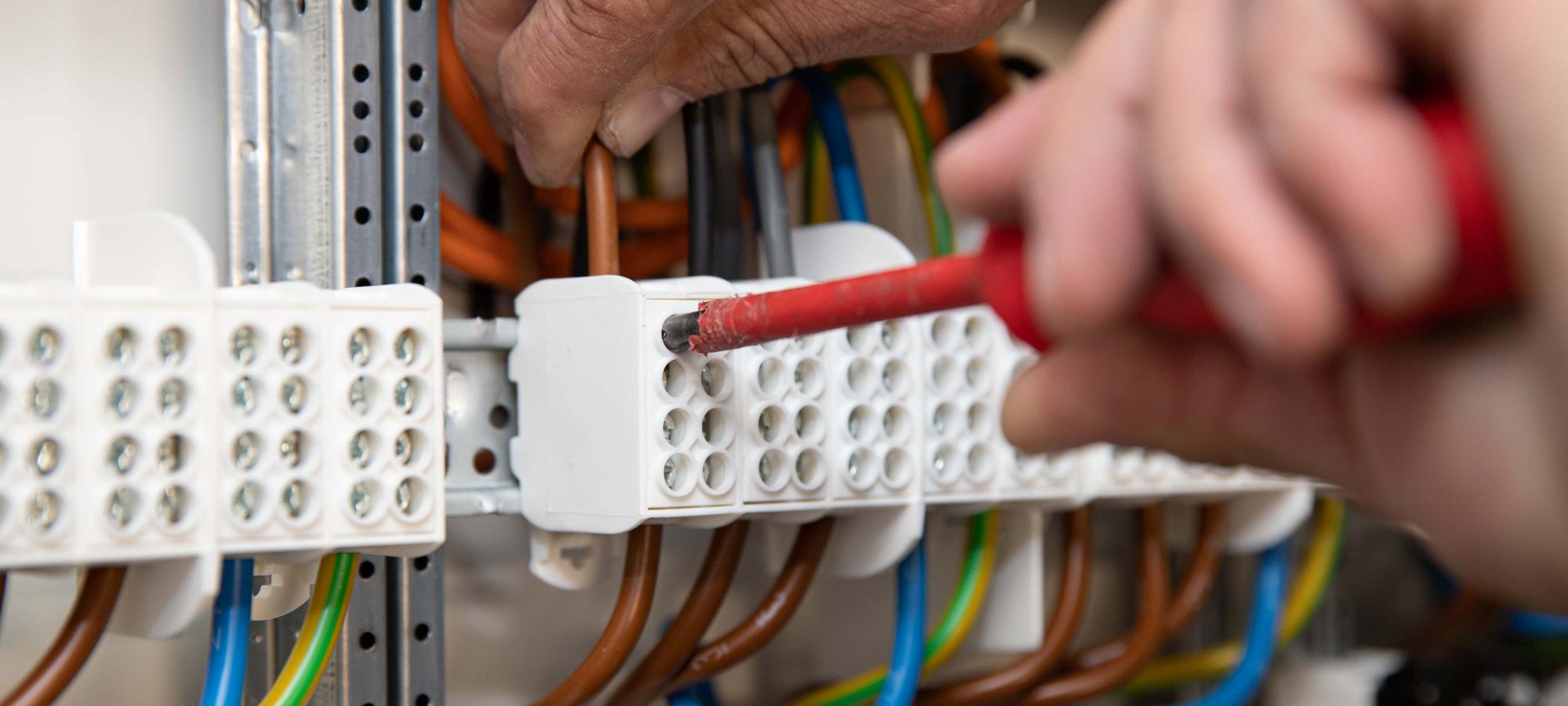 Bildausschnitt mit zwei Händen eines Elektrikermeisters - beim anschließen verschiedener Kabel mittels Schraubendrehers in einem Zählerschrank.