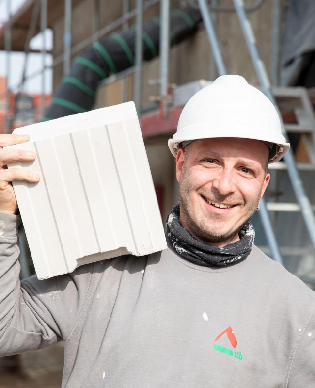 Freundlicher Bauhandwerker der ITB Dresden GmbH, mit Helm trägt einen Blockziegel auf einer Baustelle