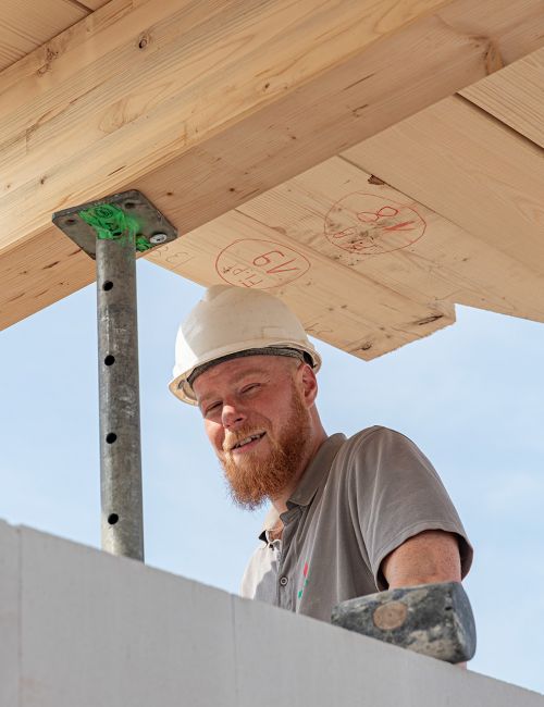 Ein fröhlicher Mitarbeiter unseres Bauhandwerkes bei Rohbauarbeiten an einem Dachgeschoss, mit Bauhelm und T-Shirt unter blauem Himmel.