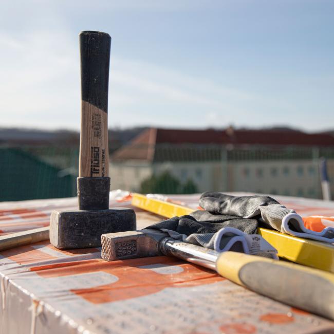 Werkzeuge - ein Hammer stehend und ein Hammer liegend, Bauhandschuhe und gelbe Wasserwaage in der Sonne liegend auf einem Stapel Ziegel einer Baustelle