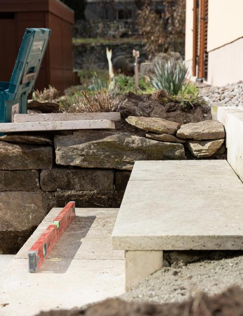 Treppen- und Terrassenbau mit hellen Natursteinplatten an einem sonnigen Tag. Ebenso zu sehen sind ein türkisfarbener Werkzeugkasten, eine orangerote Wasserwaage sowie Frühlingsblüher im einem Vorgarten mit Natursteinmauer.
