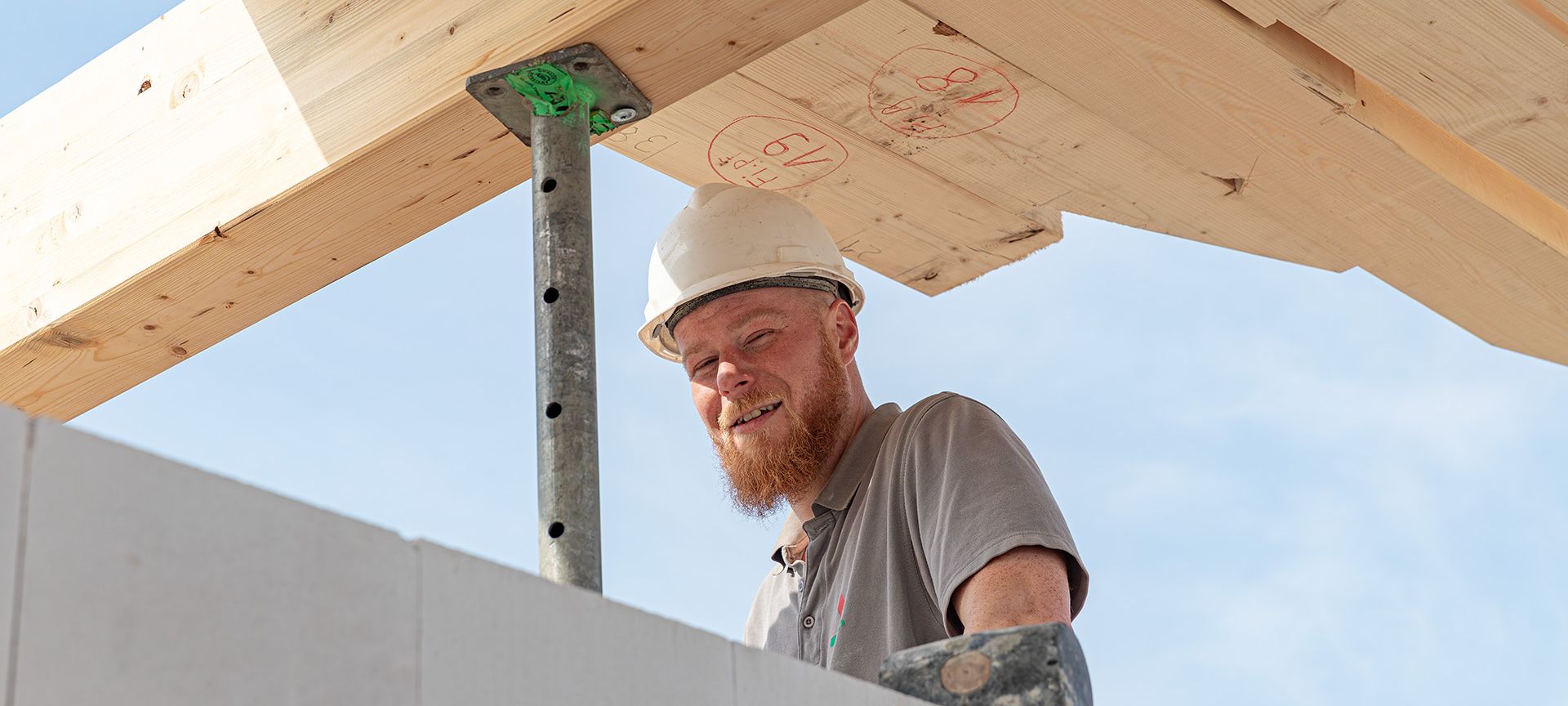 Ein fröhlicher Mitarbeiter unseres Bauhandwerkes bei Rohbauarbeiten an einem Dachgeschoss, mit Bauhelm und T-Shirt unter blauem Himmel.